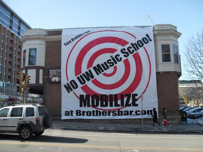 A large banner on a brick building in IL reads, "NO UW Music School, MOBILIZE at Brothersbar.com" with red target graphics. People walk by and cars, some with Car Wraps Spring Grove, are parked along the street.