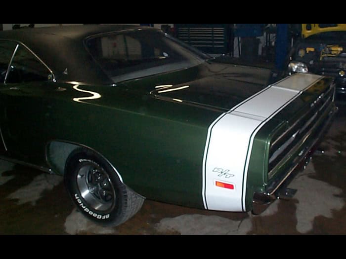 A dark green classic muscle car with a white rear stripe and “R/T” emblem, parked indoors on a wet floor. Perfect for showcasing Car Wraps Spring Grove, the car's rear quarter panel, wheel, and black vinyl roof are visible.