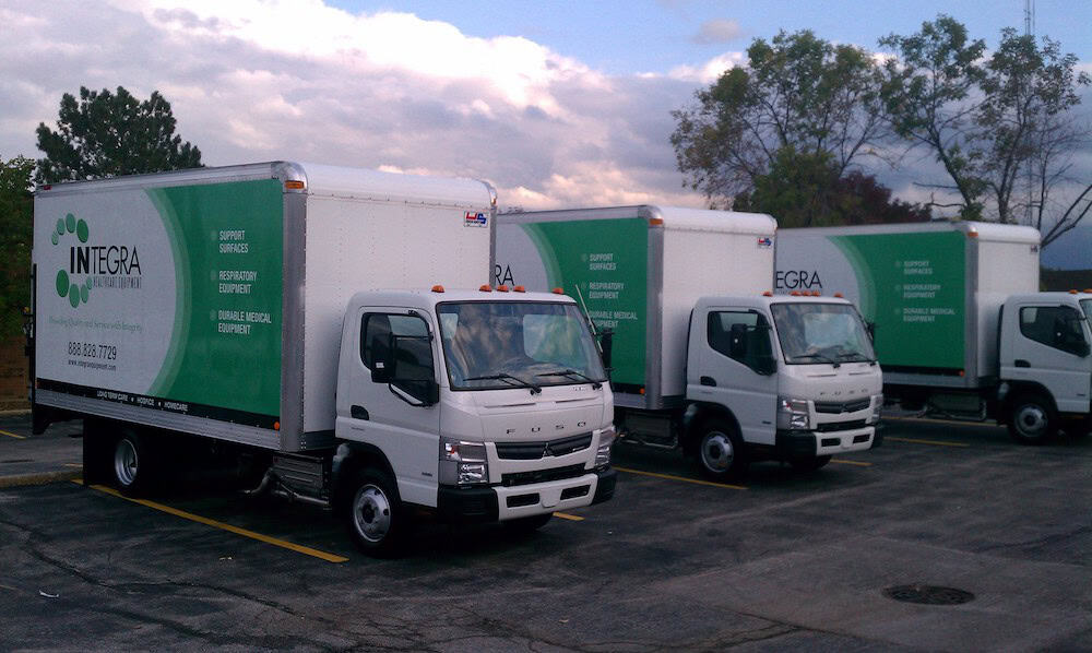 Three white box trucks with green and white "INTEGRA DELIVERY" signage, featuring vehicle wraps Spring Grove, IL, are parked side by side in a lot under a cloudy sky, with trees and a building in the background.