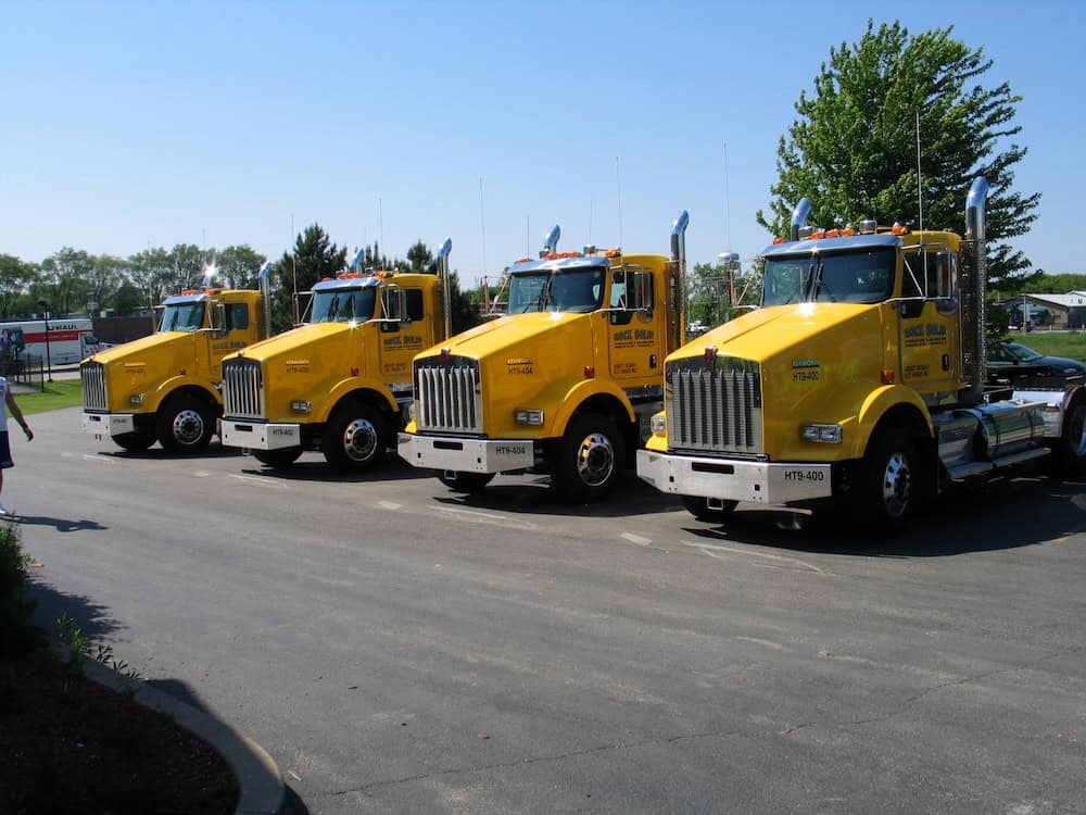 Four bright yellow semi-trucks featuring vibrant vehicle wraps Spring Grove are parked side by side on a paved lot in daylight, with green trees and a clear blue sky in the background.