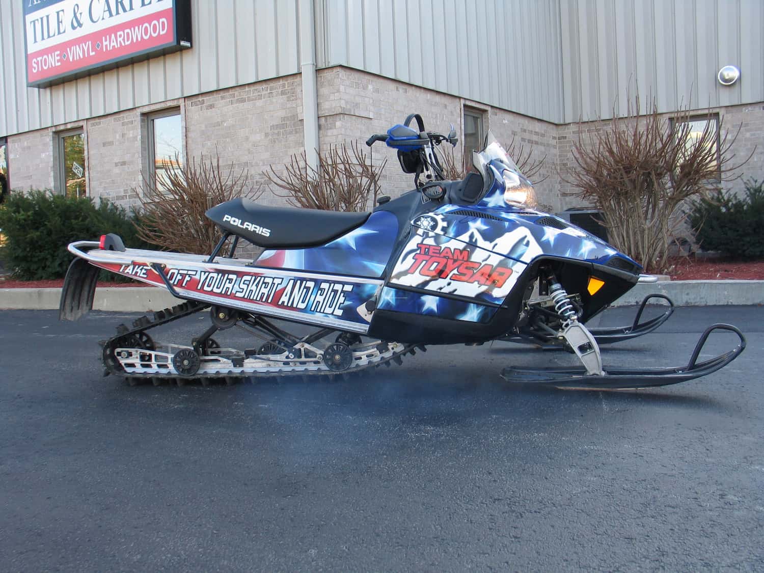 A Polaris snowmobile with "TEAM TOMSKAR" and "Take Off Your Skirt And Ride" graphics, featuring custom vehicle wraps Spring Grove, is parked on pavement in front of a building with a "Tile & Carpet" sign in IL.