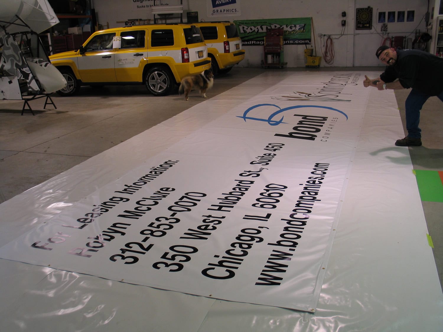 A large printed banner with leasing information and contact details lies on the floor of a workshop; a man gives a thumbs-up, while a yellow SUV—great for showcasing vehicle wraps Spring Grove—and a dog are visible in the background.