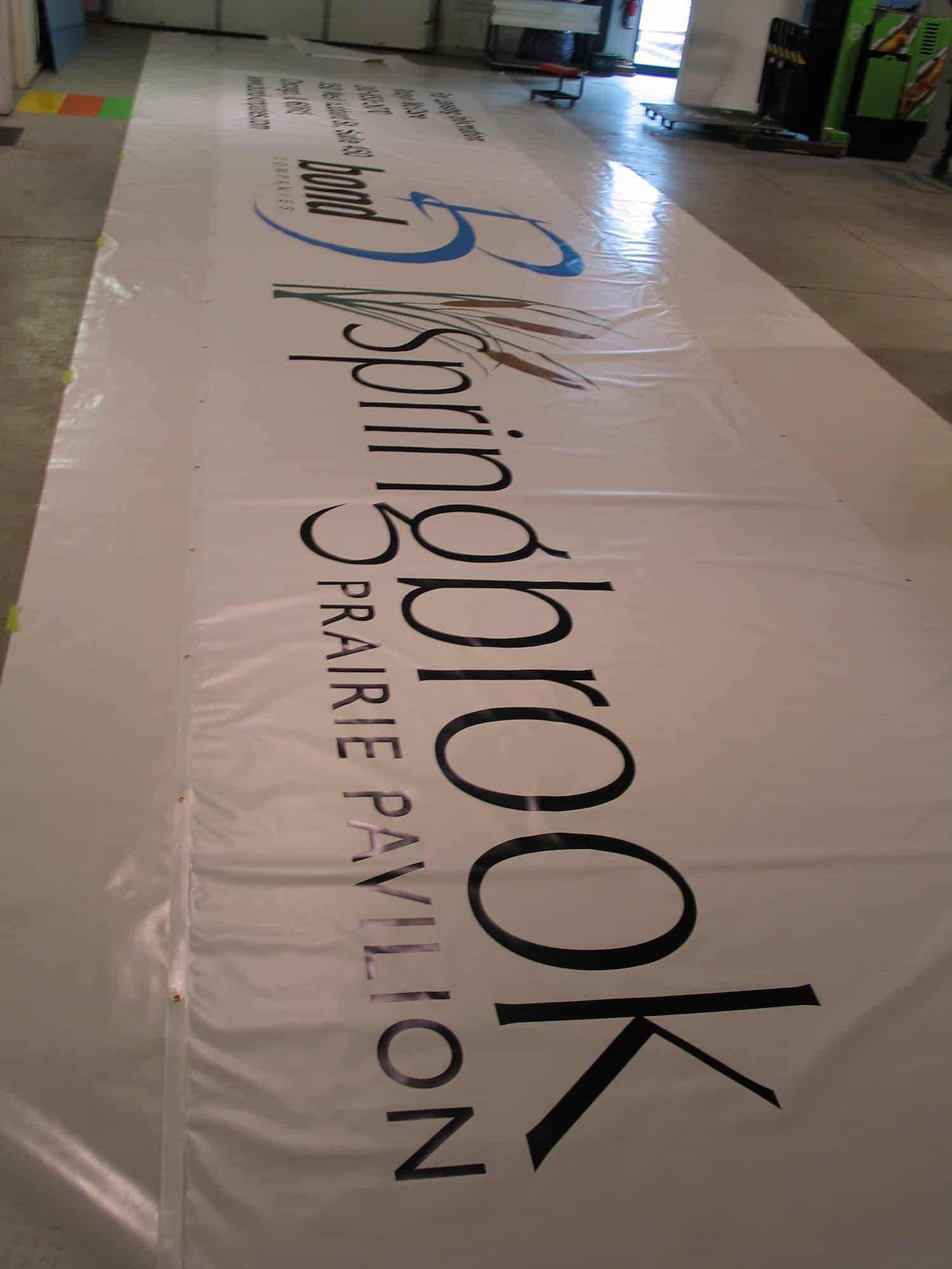 A large white banner on the floor reads “Springbrook Prairie Pavilion” with a blue and gold logo and "Naperville, IL" above the main text. The banner is laid out in a spacious indoor area.