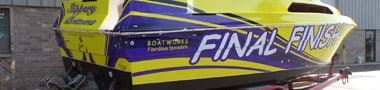 A brightly colored speedboat named "FINAL FINISH" with purple and yellow stripes, showcasing expert vehicle wraps Spring Grove, IL, is parked on a trailer outside a building. The words "Boatworks" and "Lake of the Ozarks" are also visible on the side.