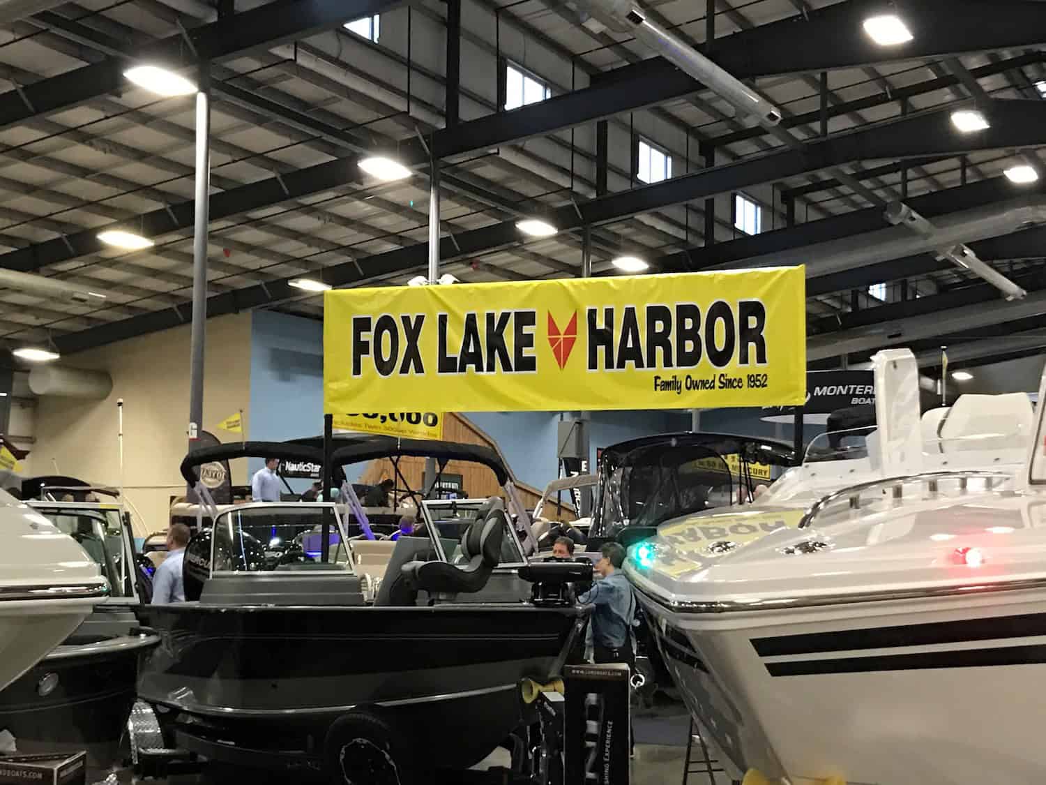 Indoor boat show with several boats on display under a large yellow banner that reads "FOX LAKE HARBOR Family Owned Since 1952." Brightly lit, with people among the boats—perfect for those interested in vehicle wraps Spring Grove events.
