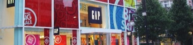 A GAP clothing store in IL with large glass windows and bold red sale signs, including “BIG SALE,” is seen from the street—trees line the right side and nearby Car Wraps Spring Grove shops add local flair.