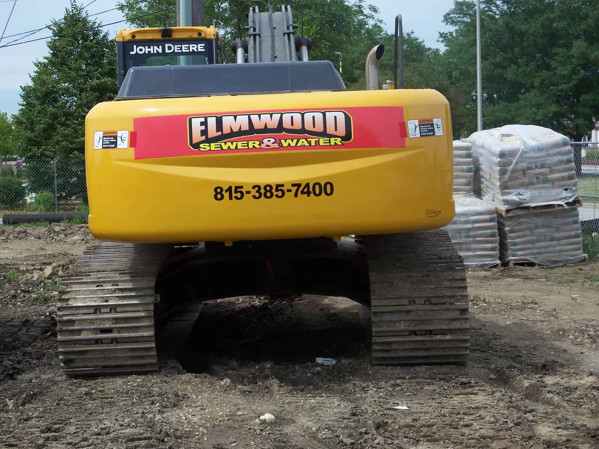 A yellow John Deere excavator labeled "ELMWOOD SEWER & WATER" with a phone number is parked on dirt, facing the camera—showcasing how vehicle wraps Spring Grove, IL can promote local businesses. Stacks of bagged materials and trees are visible in the background.