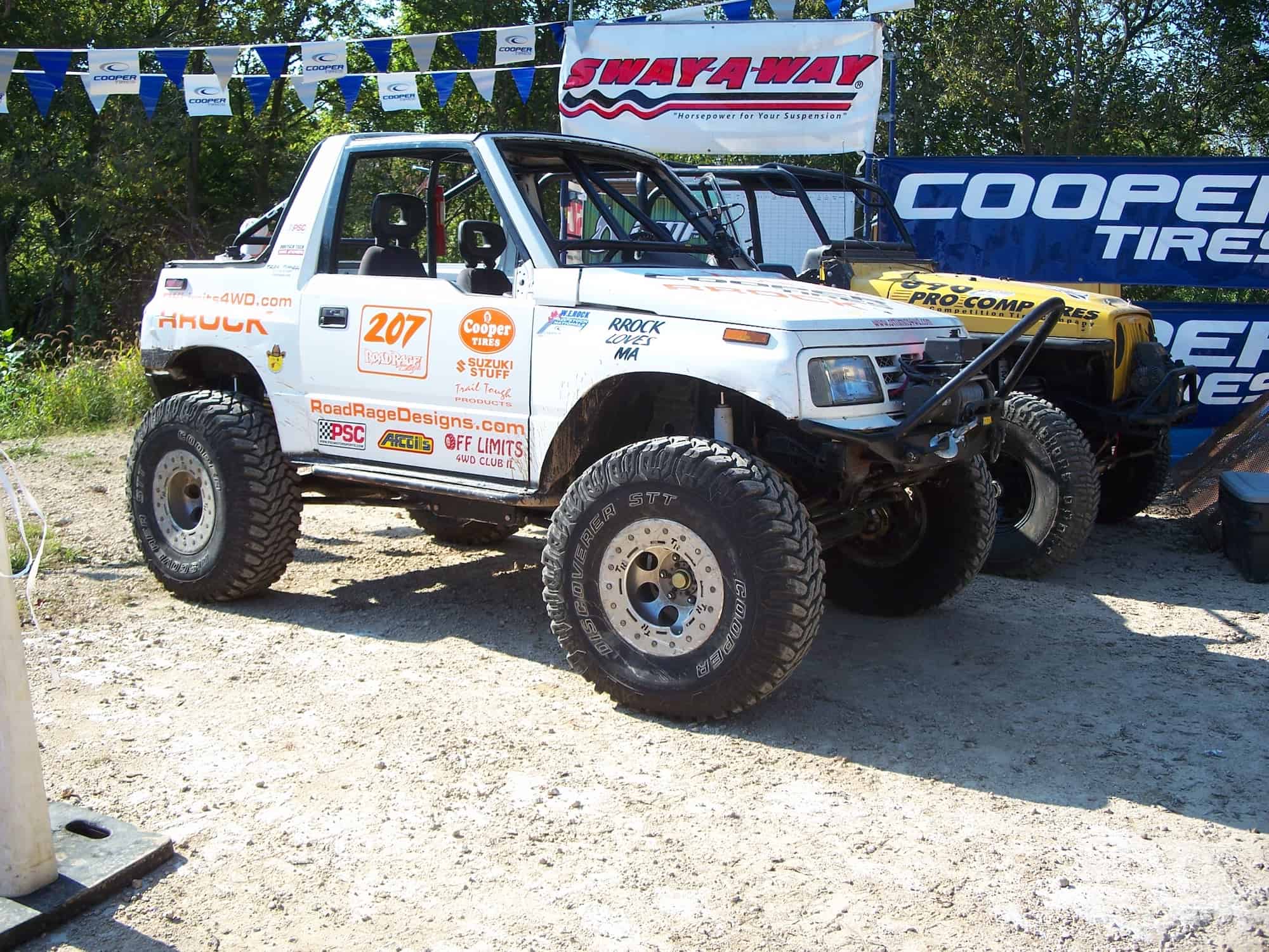 A white off-road vehicle with number 207 and various sponsor decals, showcasing Car Wraps Spring Grove, IL, is parked on a dirt surface beside a lifted yellow jeep, surrounded by banners and trees in the background.