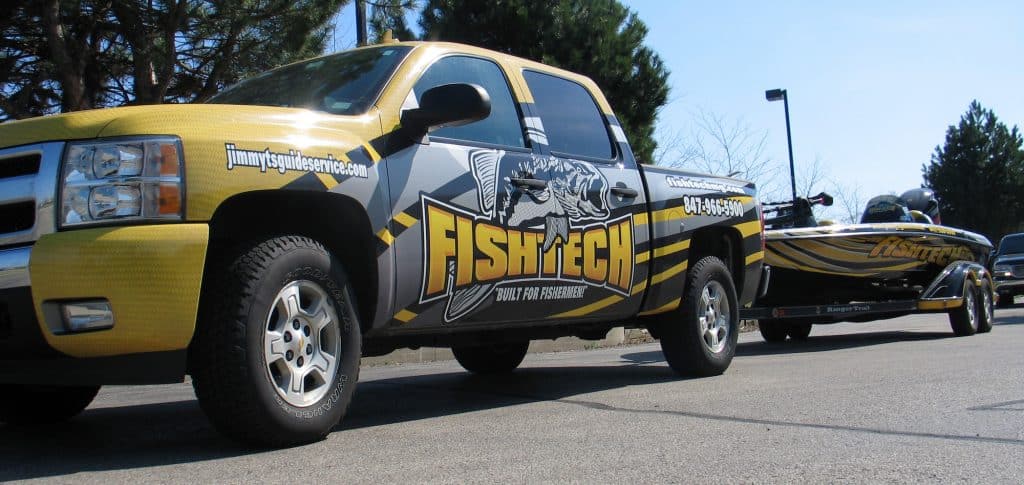A yellow and silver pickup with "Fish Tech! Built for fishermen" graphics, showcasing Car Wraps Spring Grove style, is parked on a road towing a matching fishing boat. Trees and a clear sky complete the background scene.