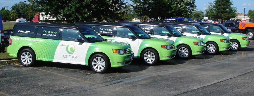 Four green and white SUVs featuring “CLEAR” branding and mobile internet ads with professional vehicle wraps Spring Grove, IL, are parked in a lot on a sunny day, surrounded by trees and other cars.