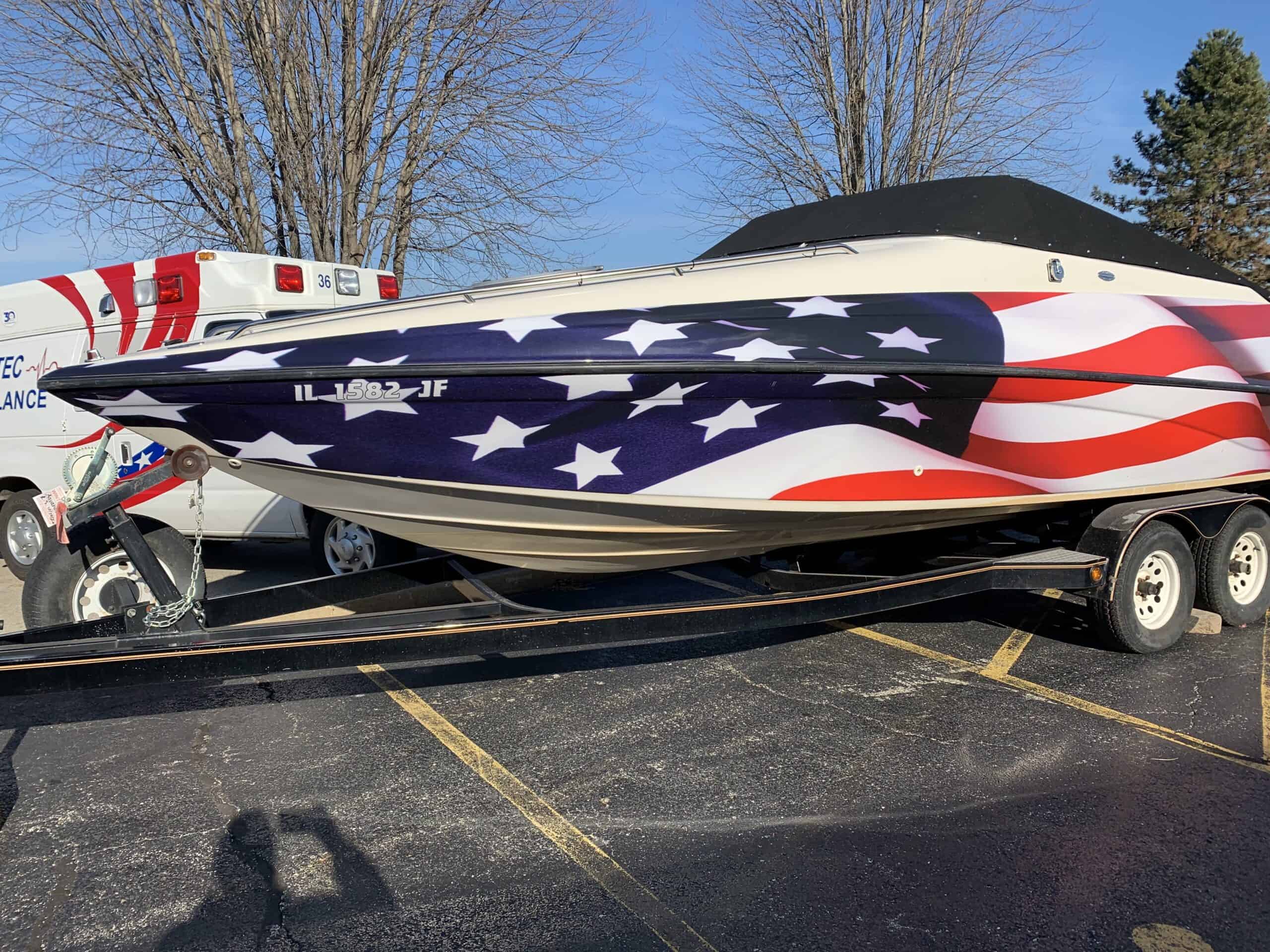 A speedboat on a trailer is painted with an American flag design, similar to the bold style of vehicle wraps Spring Grove locals love. Parked near an ambulance, it stands out against leafless trees and a clear sky in the background.