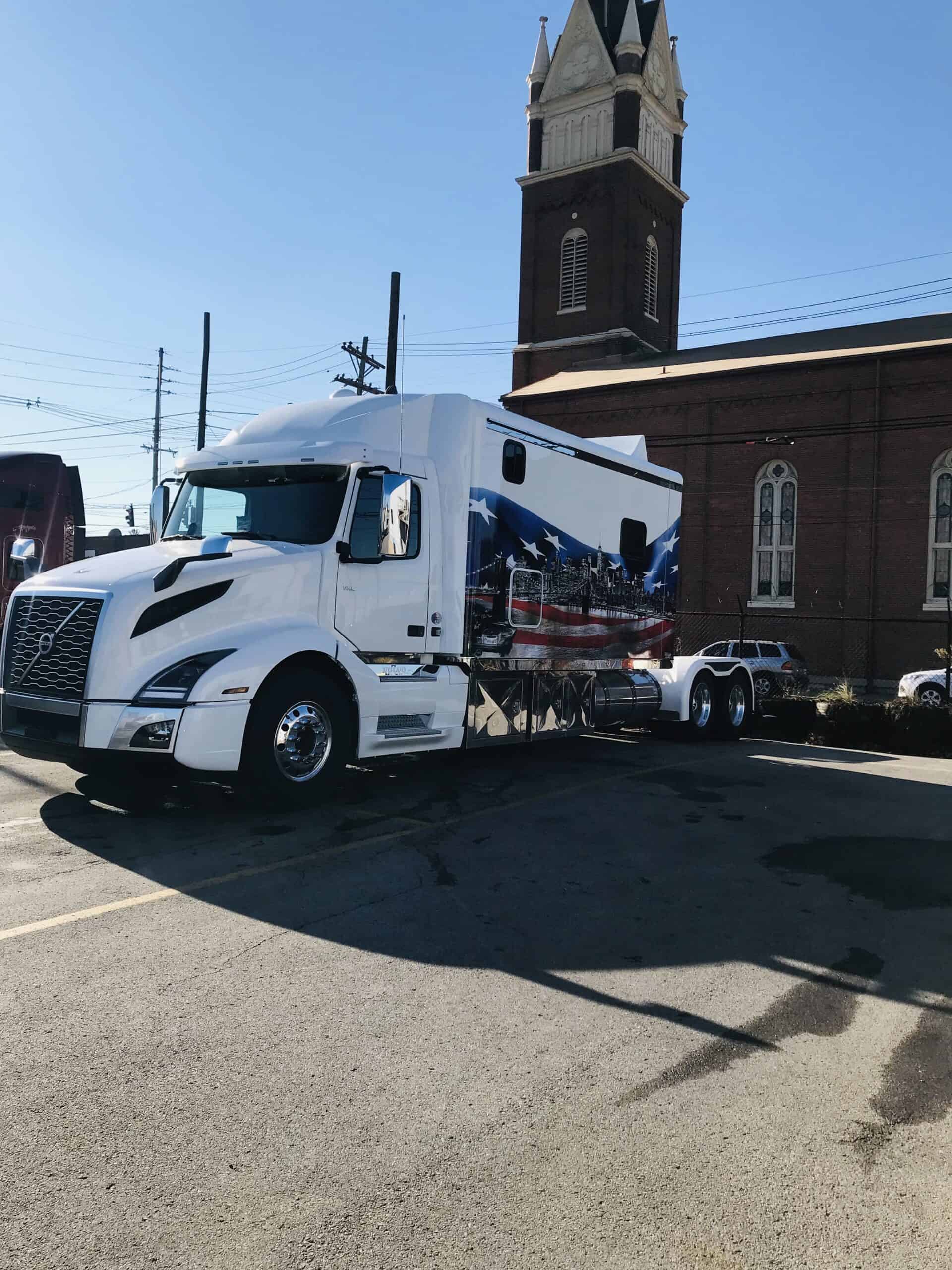 A white semi-truck with a detailed mural of the American flag and a cityscape—showcasing expert vehicle wraps Spring Grove, IL—is parked beside a brick building with tall arched windows and a church-style steeple.