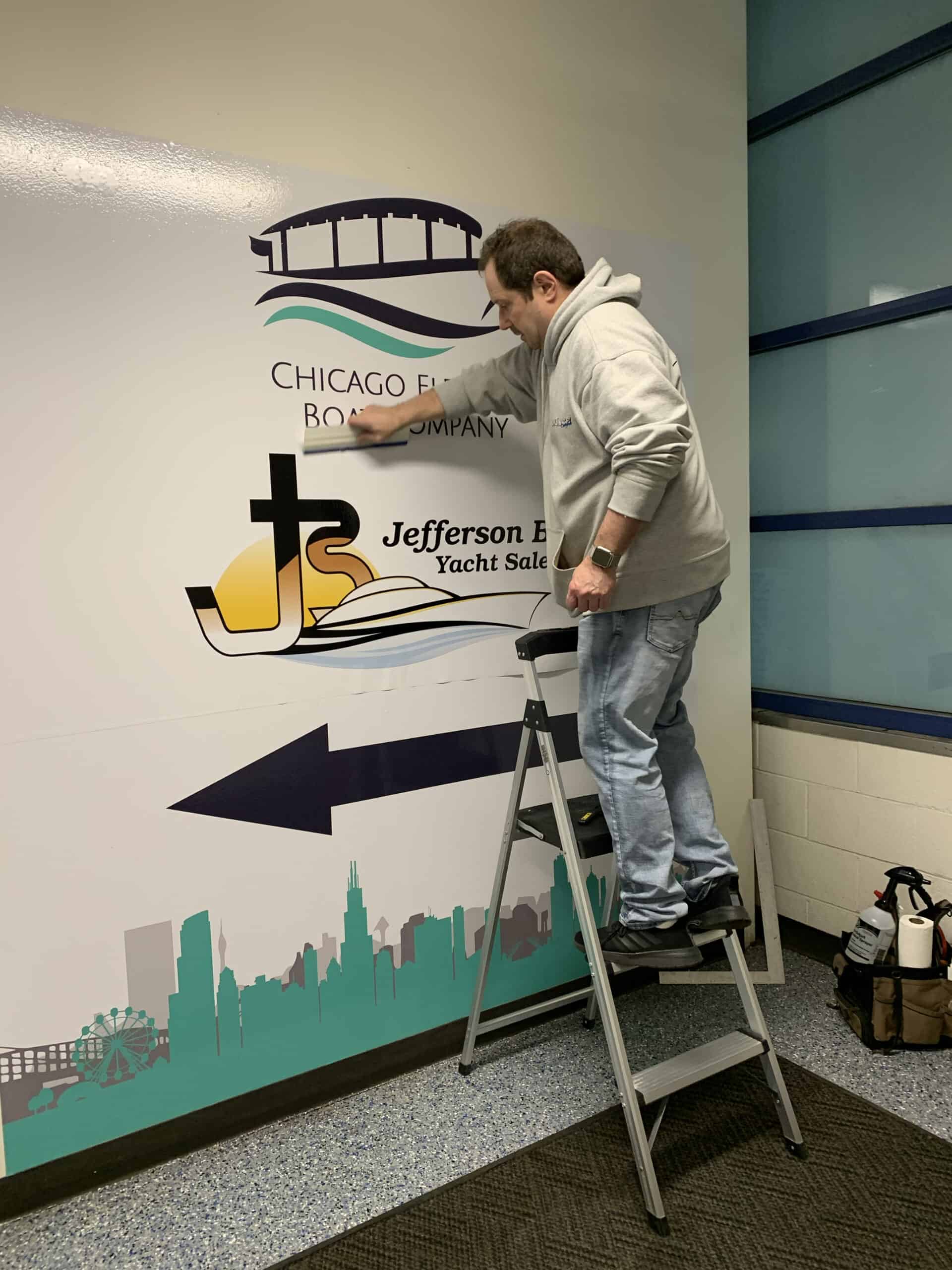 A man in a gray hoodie stands on a step ladder, applying or adjusting a wall decal for the Chicago Electric Boat Company and Jefferson Beach Yacht Sales inside a building. A bag sits on the floor nearby.