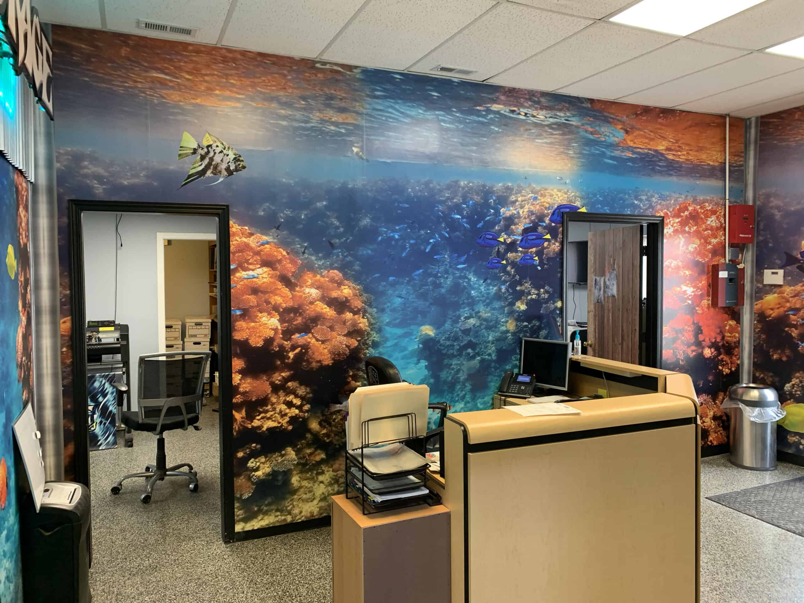 An office with desks and chairs, featuring a large, vibrant wall mural of an underwater coral reef—bringing the same creativity seen in vehicle wraps Spring Grove, IL is known for, to your workspace decor.
