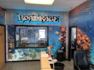 Office reception area with an underwater coral reef wall mural, a black chair, desk, and a window showing another room. Above the window is a large sign reading "ROAD RAGE" with a blue glow—home of expert Car Wraps Spring Grove, IL.
