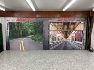 A wall display features two large photos: a winding road through a green forest and an urban street scene, both branded with "Nielsen Enterprises," specialists in vehicle wraps Spring Grove and Car Wraps Spring Grove, IL.