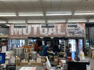 A hardware store entrance in IL with a large sign reading “MUTUAL” above the door, brick wall design, vintage truck graphics like those seen on vehicle wraps Spring Grove, and “Since 1917.” Store shelves with assorted products are visible in the foreground.