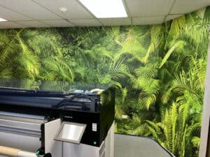 A large-format printer stands in front of a wall covered with vibrant, green tropical leaf wallpaper, creating a lush jungle-like atmosphere—perfect for designing custom vehicle wraps Spring Grove, IL.