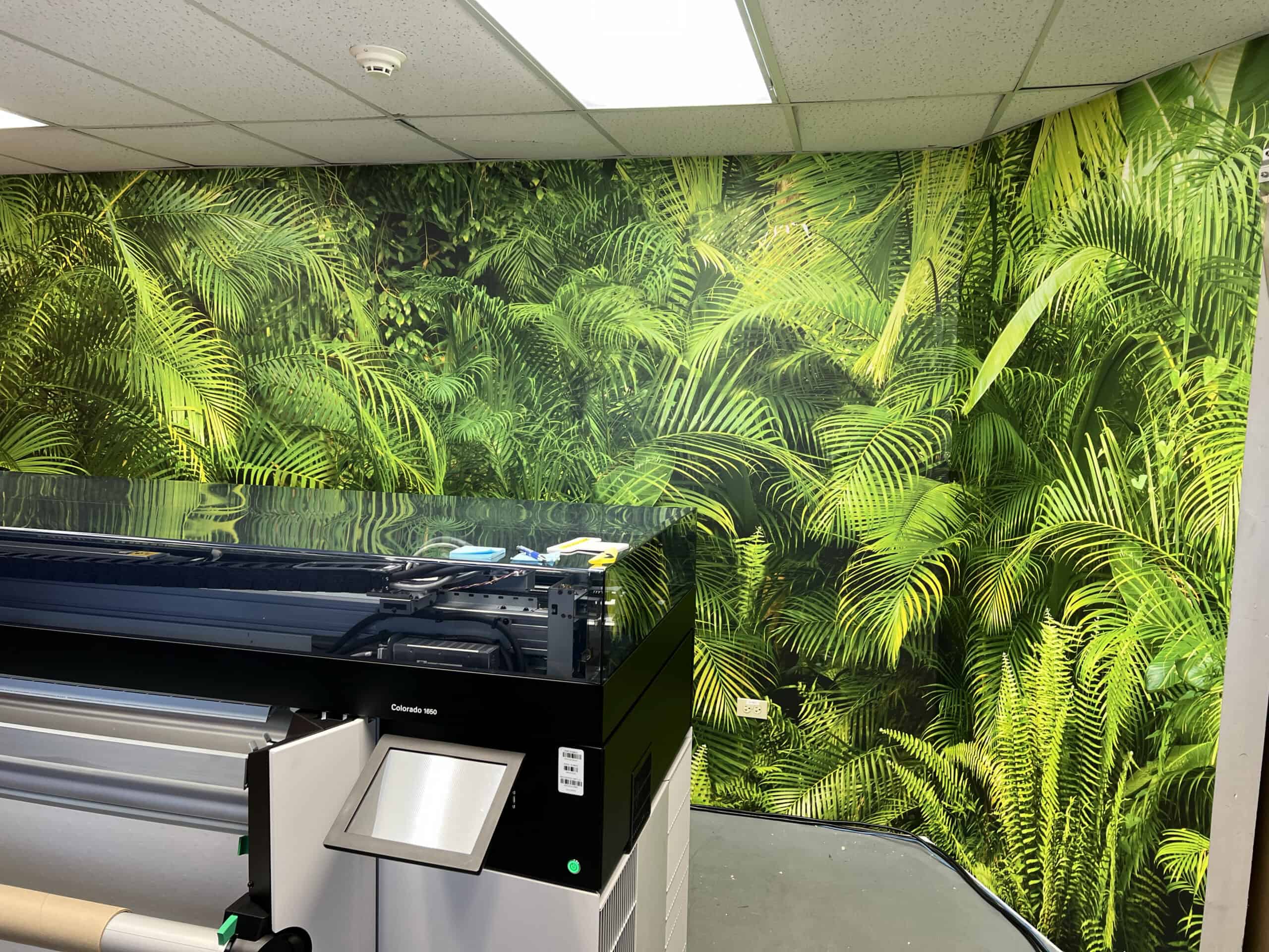 A large-format printer stands in front of a wall covered with vibrant, green tropical leaf wallpaper, creating a lush jungle-like atmosphere—perfect for designing custom vehicle wraps Spring Grove, IL.