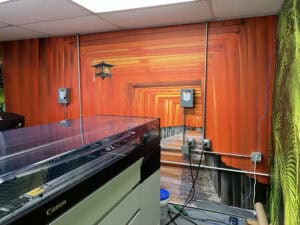 A printer sits in a room with a vibrant wall mural depicting a pathway lined with orange torii gates, reminiscent of the Fushimi Inari Shrine in Japan. Electrical boxes and cables are mounted on the mural wall, reflecting creative vibes found at Car Wraps Spring Grove, IL.