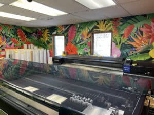 A large printer sits in a vibrant room with tropical floral wallpaper, two windows, and rolls of blank paper or vinyl stacked in the corner—the perfect setting for crafting vehicle wraps Spring Grove studios in IL love.