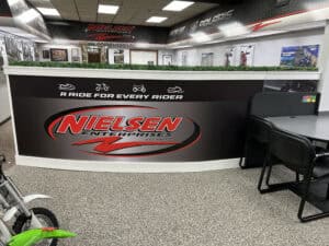 A modern showroom in IL with a large Nielsen Enterprises sign on the counter reading “A ride for every rider.” Chairs, tables, framed pictures, and a green artificial plant create a welcoming space for vehicle wraps Spring Grove customers.