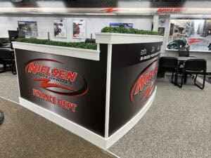 A modern office area features a black counter labeled "Nielsen Enterprises Finance Dept" with red and white branding, fake greenery on top, and chairs and posters in the background—showcasing expertise in vehicle wraps Spring Grove.