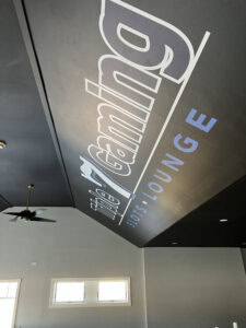 Large wall text reads "Triple 7 Gaming Slots • Lounge" in bold white and blue letters on a dark wall. Ceiling fan and three rectangular windows are visible, with sunlight streaming into the modern room—perfect for discussing vehicle wraps Spring Grove, IL.