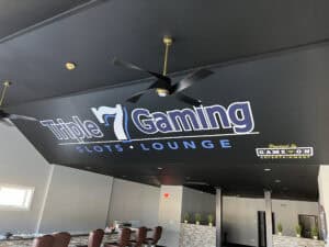Interior of a gaming lounge with "Triple 7 Gaming Slots Lounge" and "Powered by GameOn Entertainment" logos on a black ceiling, plus ceiling fans and seating areas below. Ask about Car Wraps Spring Grove for custom vehicle wraps in IL.