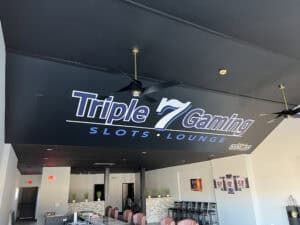 Interior of a gaming lounge in IL with a black ceiling featuring large "Triple 7 Gaming Slots Lounge" signage, ceiling fans, a bar area with pink chairs, and unique decor like plants and framed jerseys—perfect after checking out car wraps Spring Grove offers nearby.