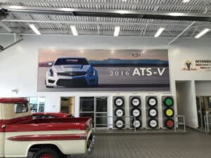 Large wall banner inside a car dealership displays a Cadillac ATS-V with "First Ever 2016 ATS-V" text. Below are glass doors, pricing signs, and part of a classic red and white pickup—perfect inspiration for vehicle wraps Spring Grove enthusiasts.