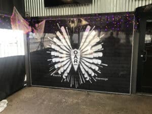 A black wall mural features white painted wings made of oars with the OAR logo at the center. Purple string lights frame the mural, and hashtags #oarlife and #oarwings are written below—crafted by experts in vehicle wraps Spring Grove.