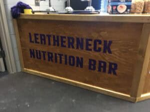 A wooden counter with the words "LEATHERNECK NUTRITION BAR" painted in purple letters. Some food items and equipment are visible on top, giving a local vibe like shops offering vehicle wraps Spring Grove residents trust.