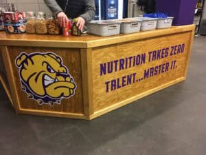 A wooden snack bar in Spring Grove, IL, with a fierce bulldog logo and “NUTRITION TAKES ZERO TALENT… MASTER IT.” A person arranges snacks on the counter, while baskets of food are displayed—like a vehicle wrap for your taste buds!.