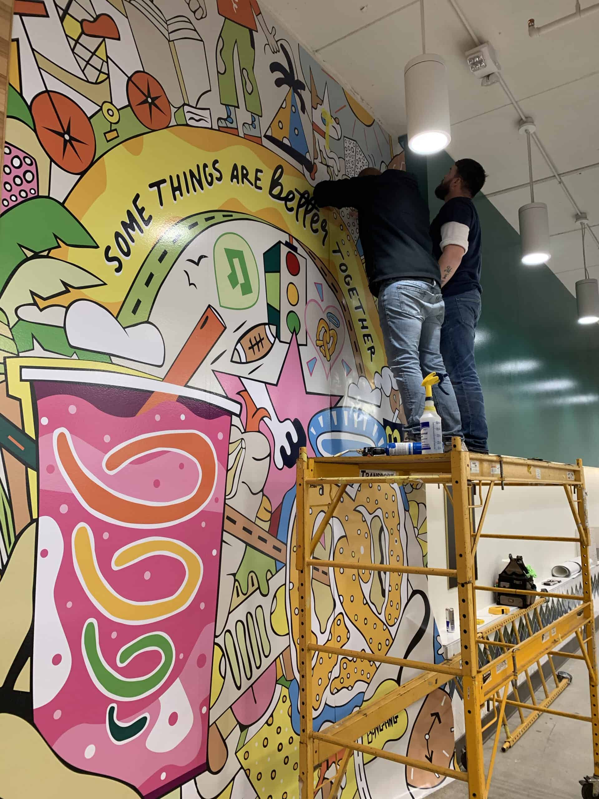 Two people on scaffolding work on a vibrant mural with food, drinks, and the phrase "Some things are better together" in a bright indoor space—similar teamwork goes into creating stunning Car Wraps Spring Grove projects.