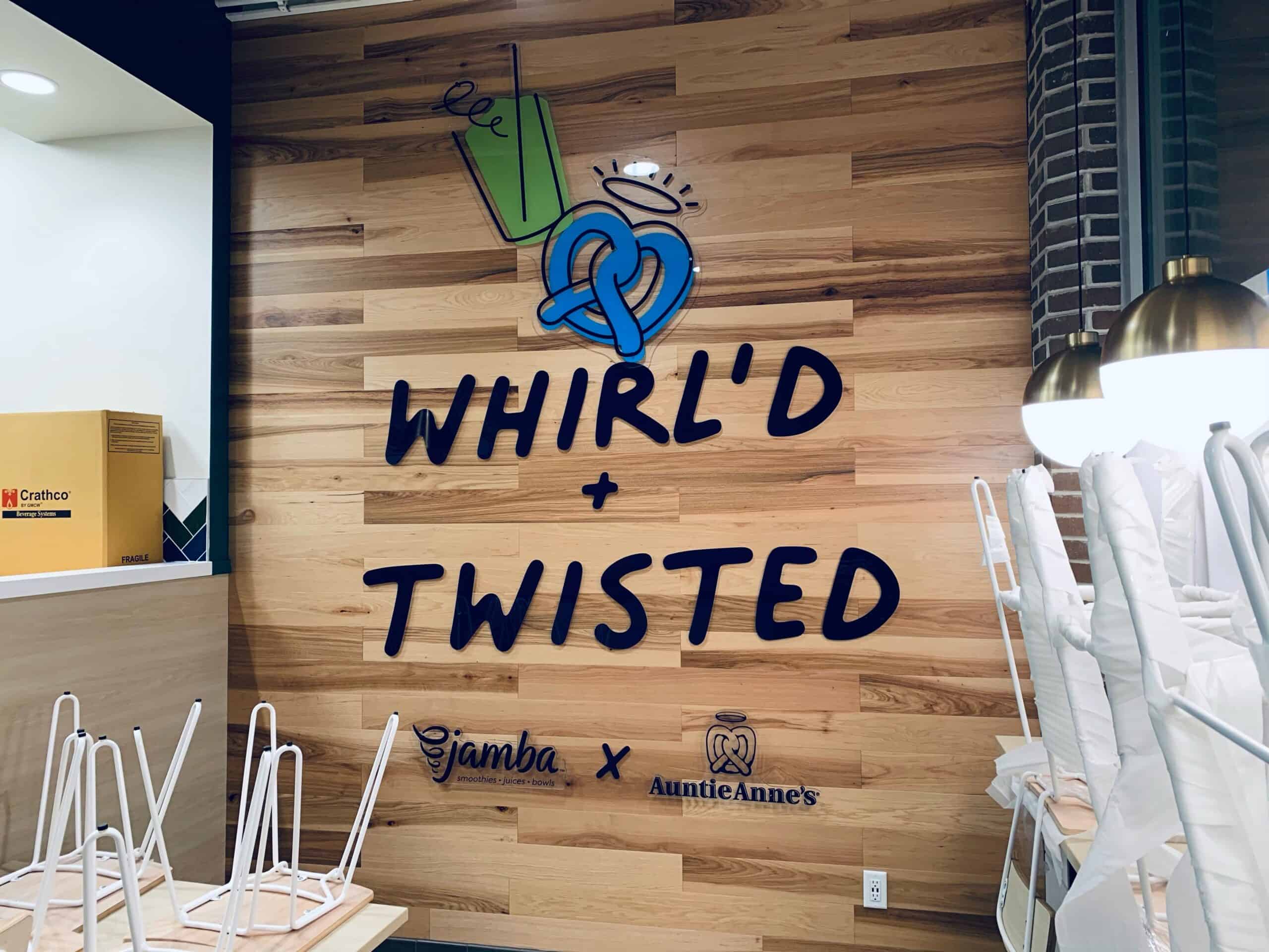 A wooden wall features a blue pretzel and cup graphic above the words “WHIRL’D + TWISTED,” with Jamba and Auntie Anne’s logos below; stacked chairs are visible in the foreground—like custom Car Wraps Spring Grove.