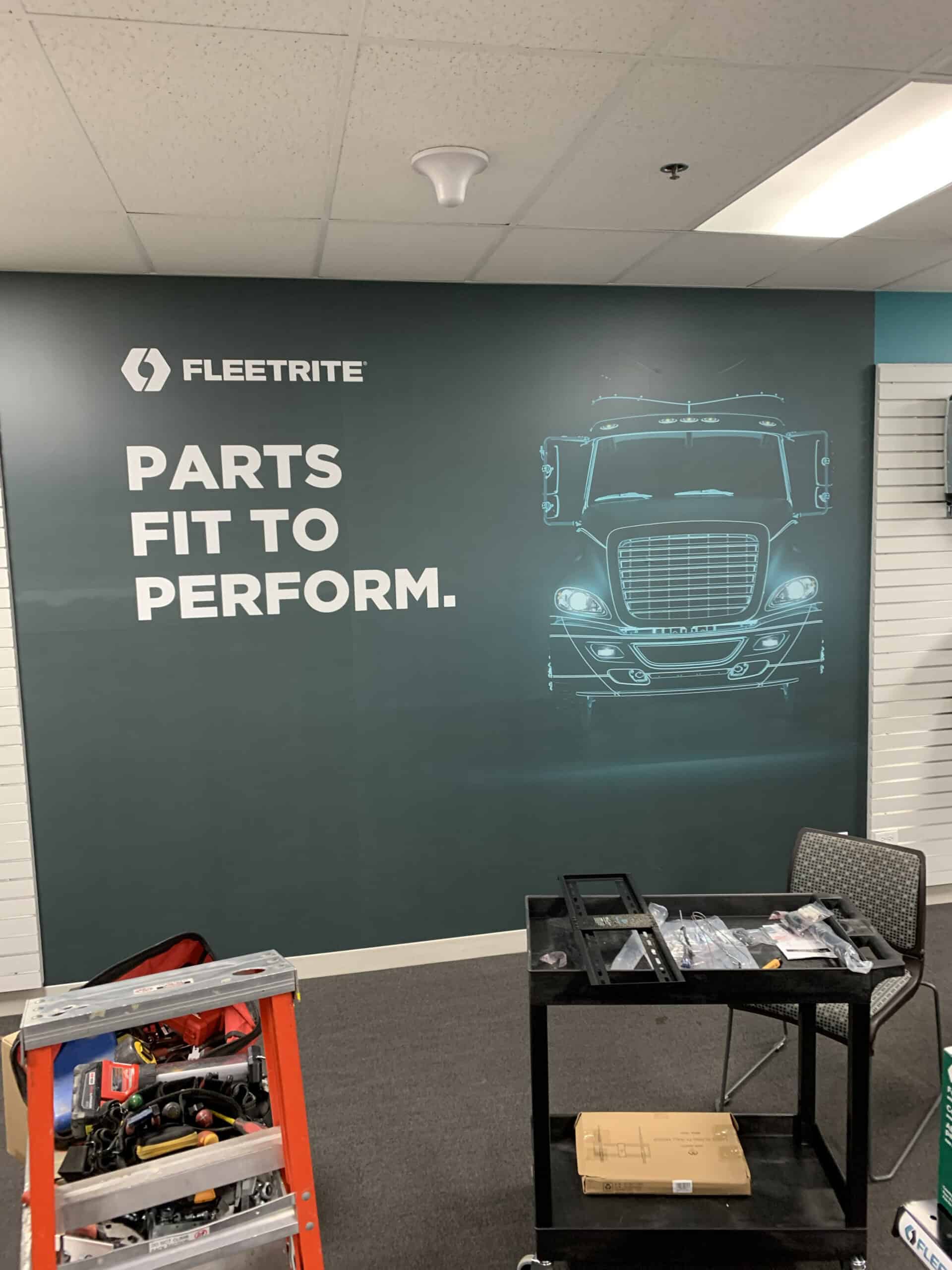 A wall display shows the FLEETRITE logo, "PARTS FIT TO PERFORM," and an outlined truck front. In front, a tool case, orange ladder, chair, and various tools are visible—perfect for those seeking vehicle wraps Spring Grove, IL.