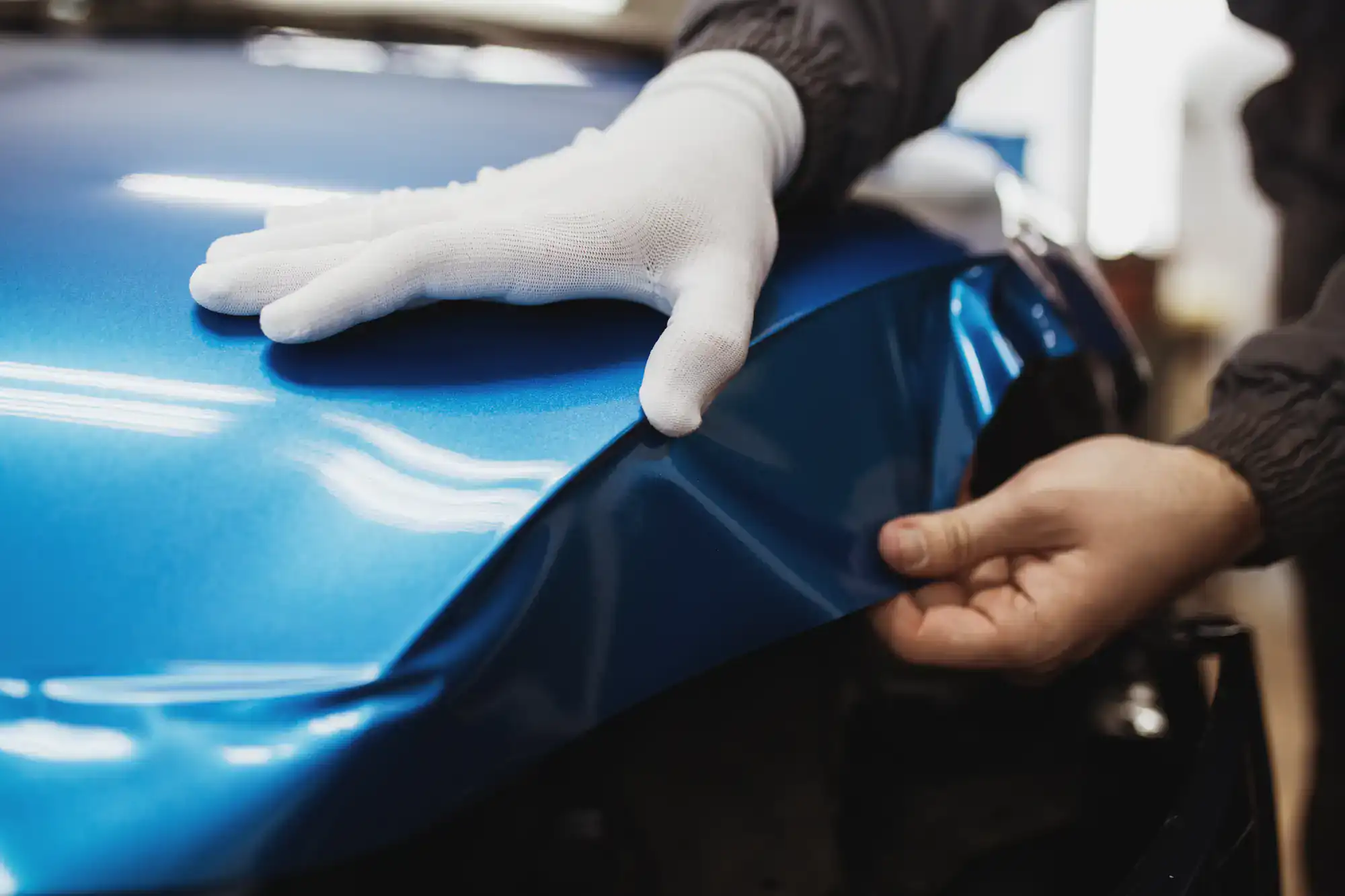 A person wearing white gloves applies a blue vinyl wrap to the hood of a car in Spring Grove, IL, smoothing the material for proper adhesion and a flawless finish—showcasing expert vehicle wraps Spring Grove craftsmanship.