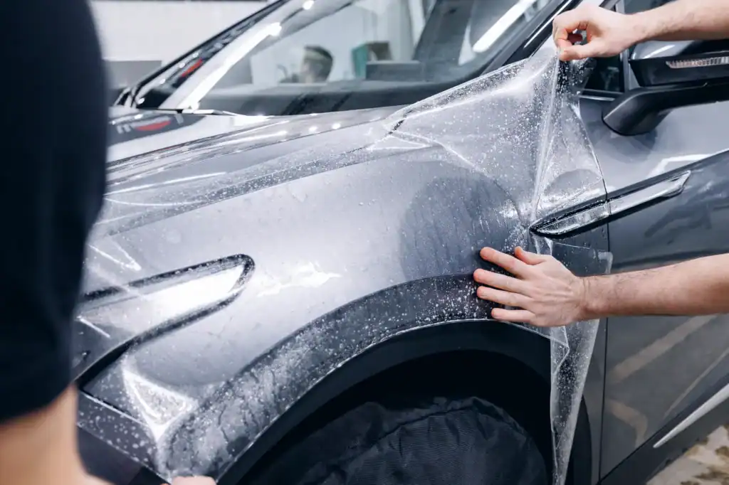 Two people in IL apply a clear protective film to the front fender of a gray car, smoothing it with their hands to prevent bubbles and ensure full coverage, showcasing expert Car Wraps Spring Grove services.