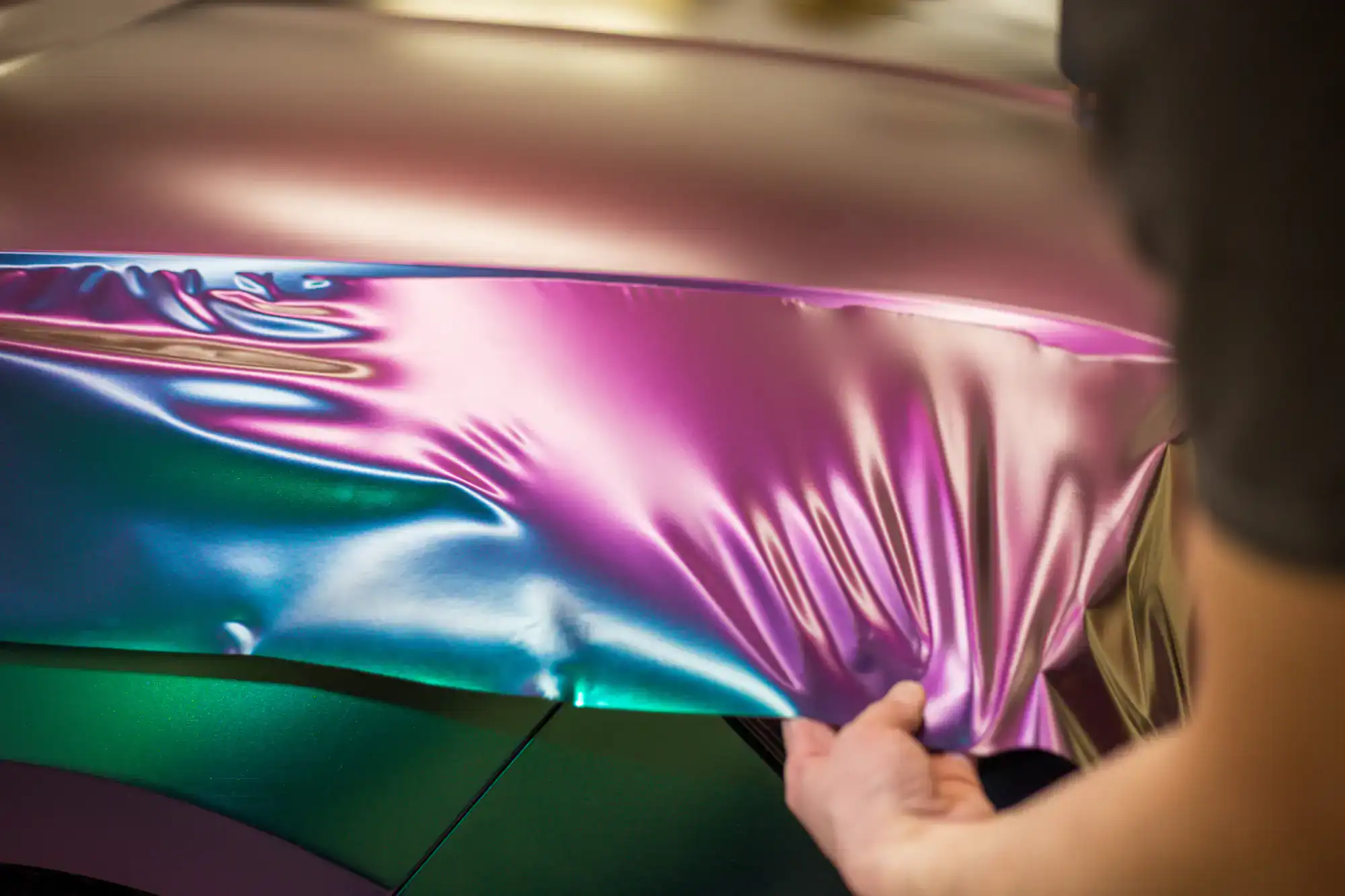A person applies a glossy, iridescent vinyl wrap in pink, purple, and blue shades to the side of a car, smoothing the material by hand—showcasing expert Car Wraps Spring Grove craftsmanship.