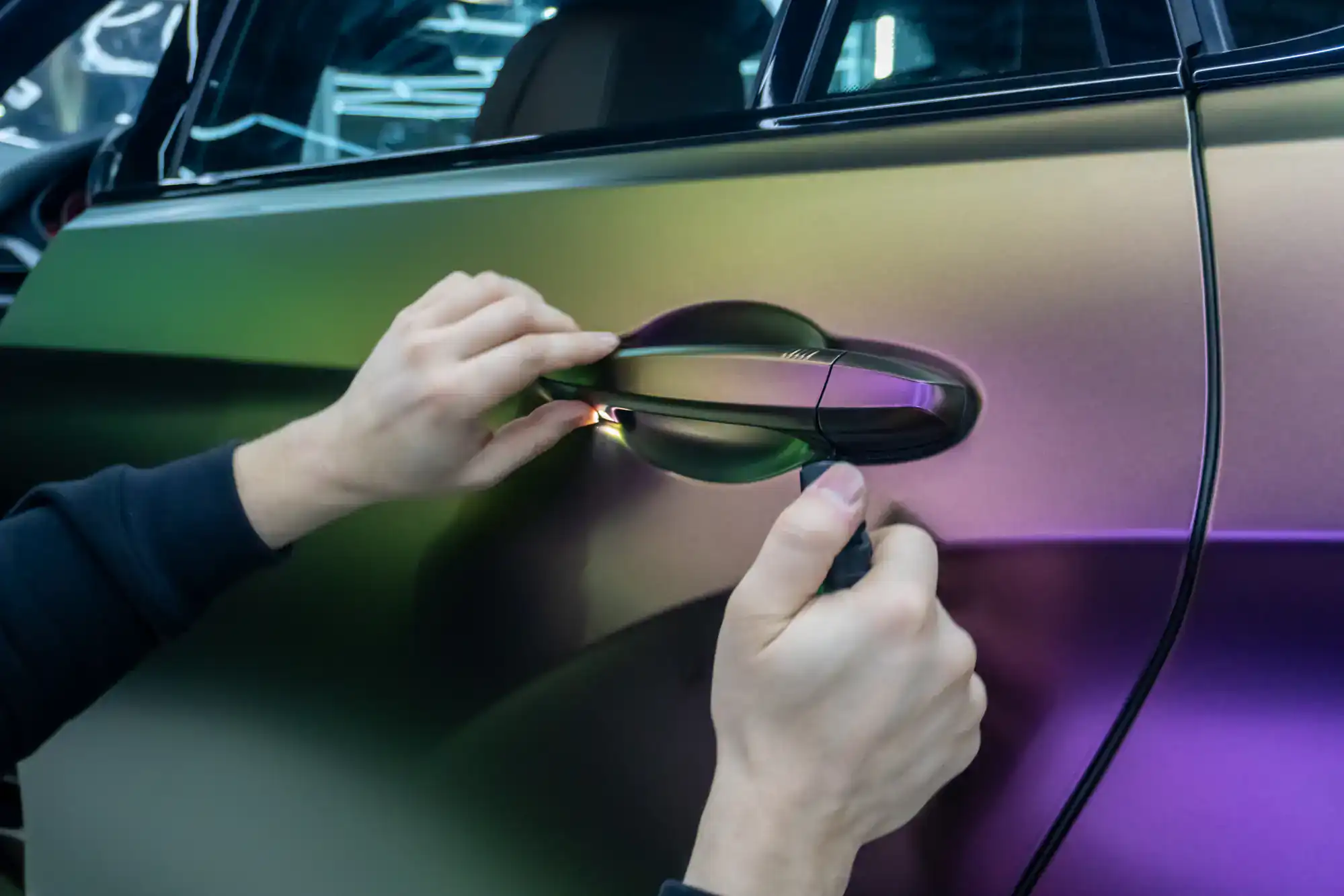 A person uses a tool to carefully install or wrap a car door handle on a vehicle with a shiny, iridescent paint finish—showcasing expert Car Wraps Spring Grove, IL craftsmanship.