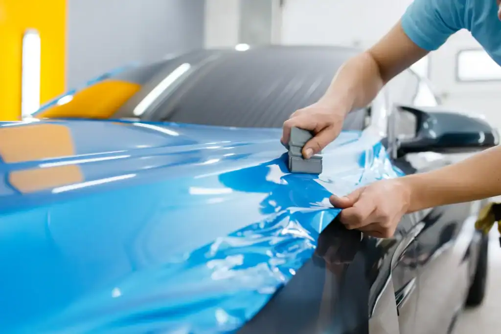 A person applies a blue vinyl wrap to the hood of a car, smoothing the material with a tool to ensure a seamless finish in an automotive workshop.