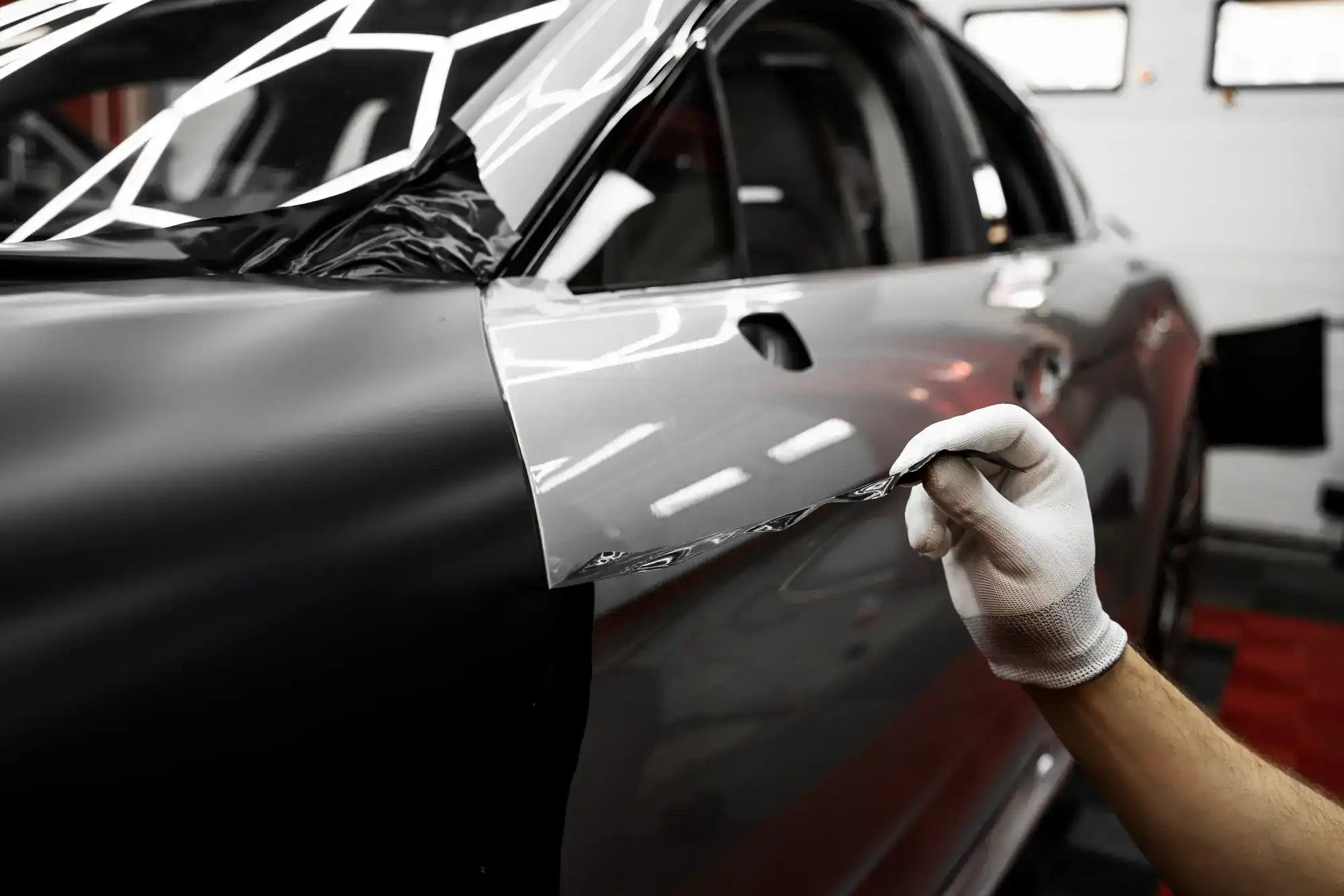 A gloved hand applies a vinyl wrap to the side of a black car, partially covering the door, in a well-lit garage or workshop.