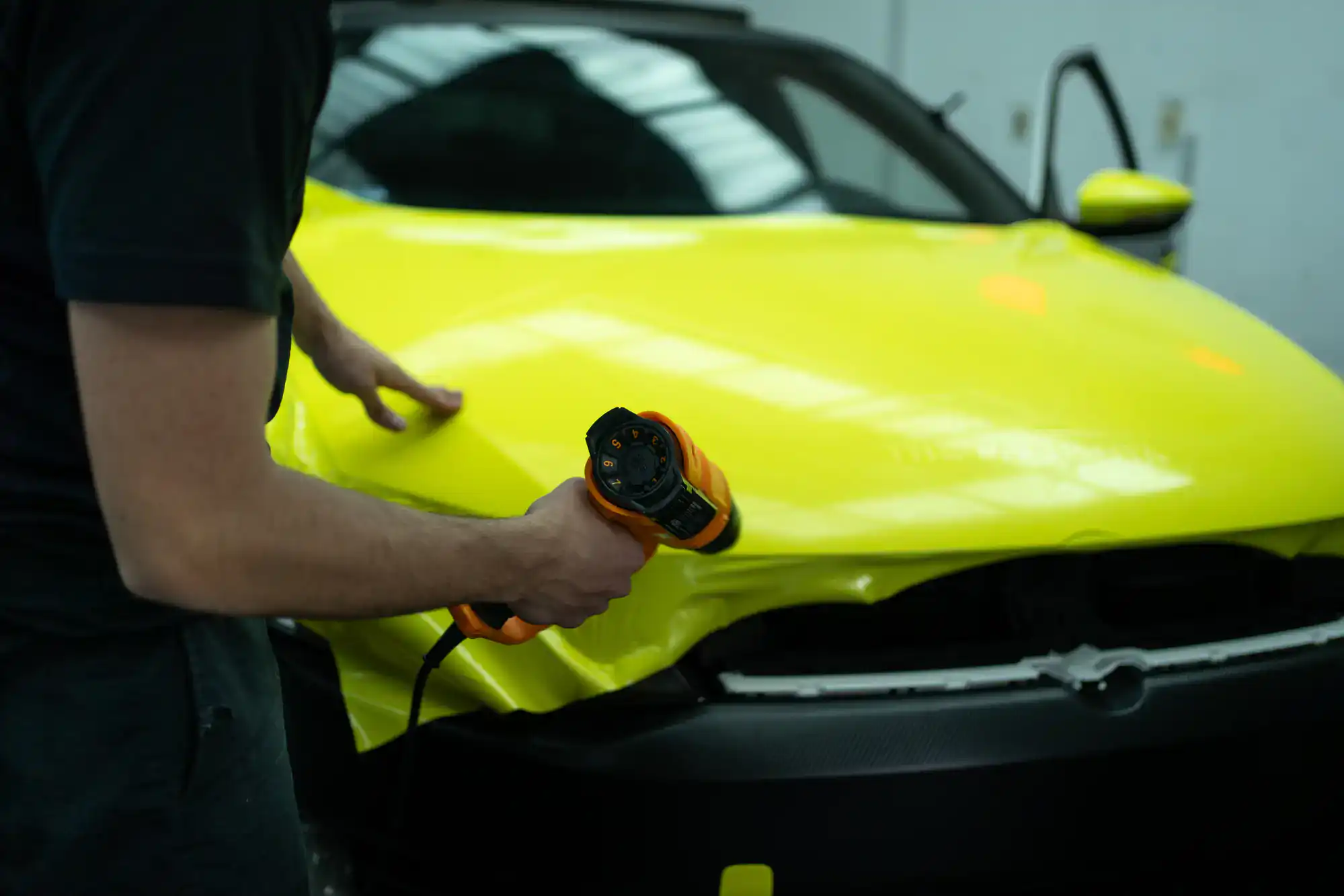 A person uses a heat gun to apply a bright yellow vinyl wrap to the hood of a car in a workshop. The car's front grille and one door are visible.