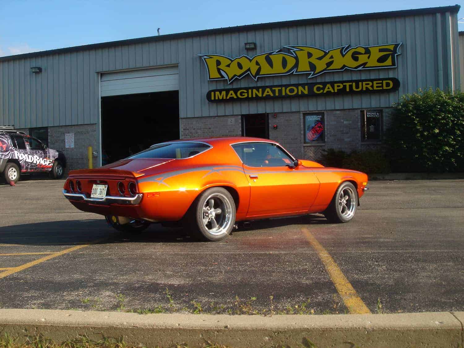 A shiny orange classic muscle car is parked in front of a building with a sign that reads "Road Rage Imagination Captured." Another similar vehicle is partially visible on the left.