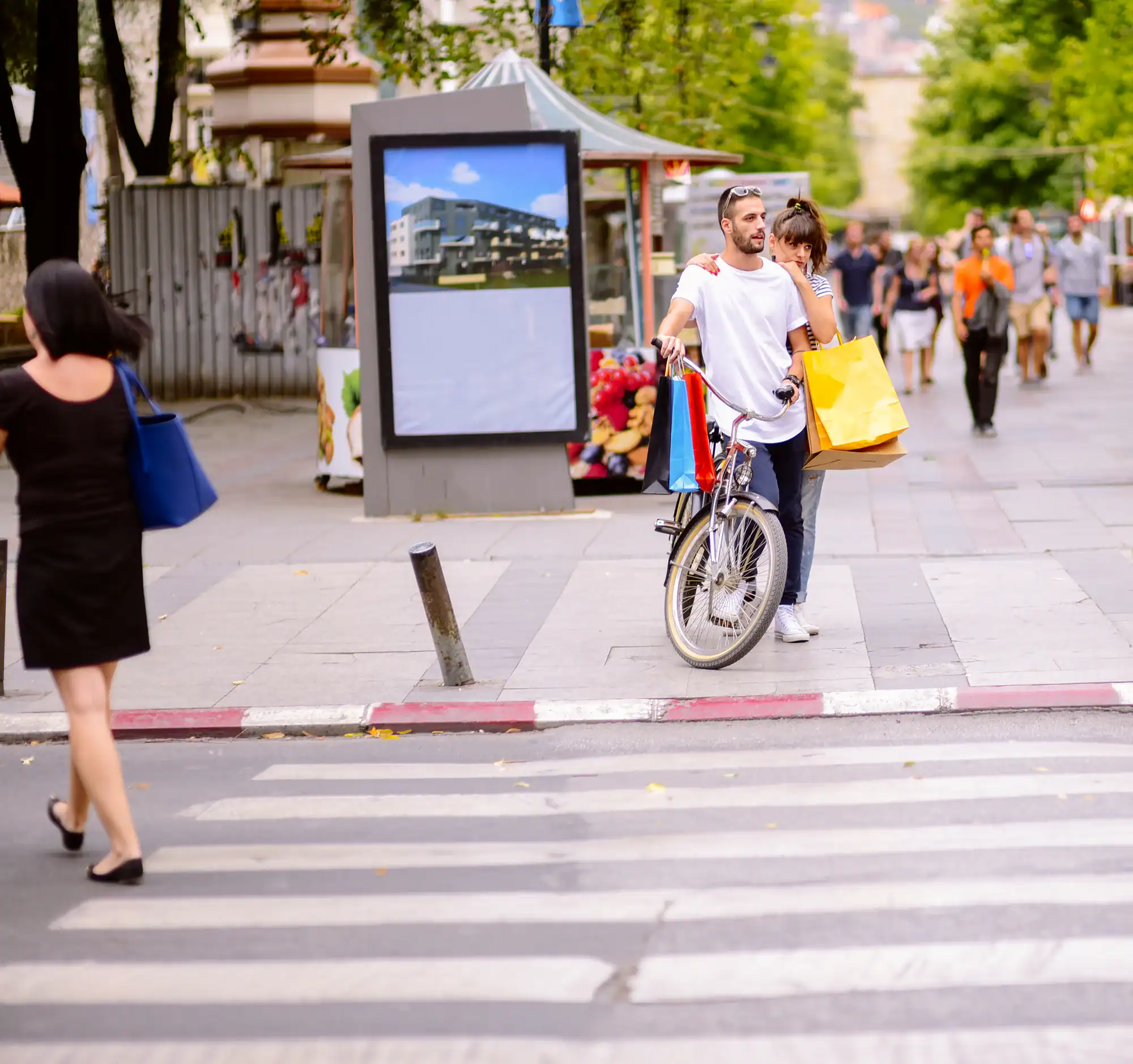 A man with a bicycle and a woman carrying shopping bags stand at a crosswalk on a city street. Other pedestrians are walking nearby, and there is a billboard and trees in the background.