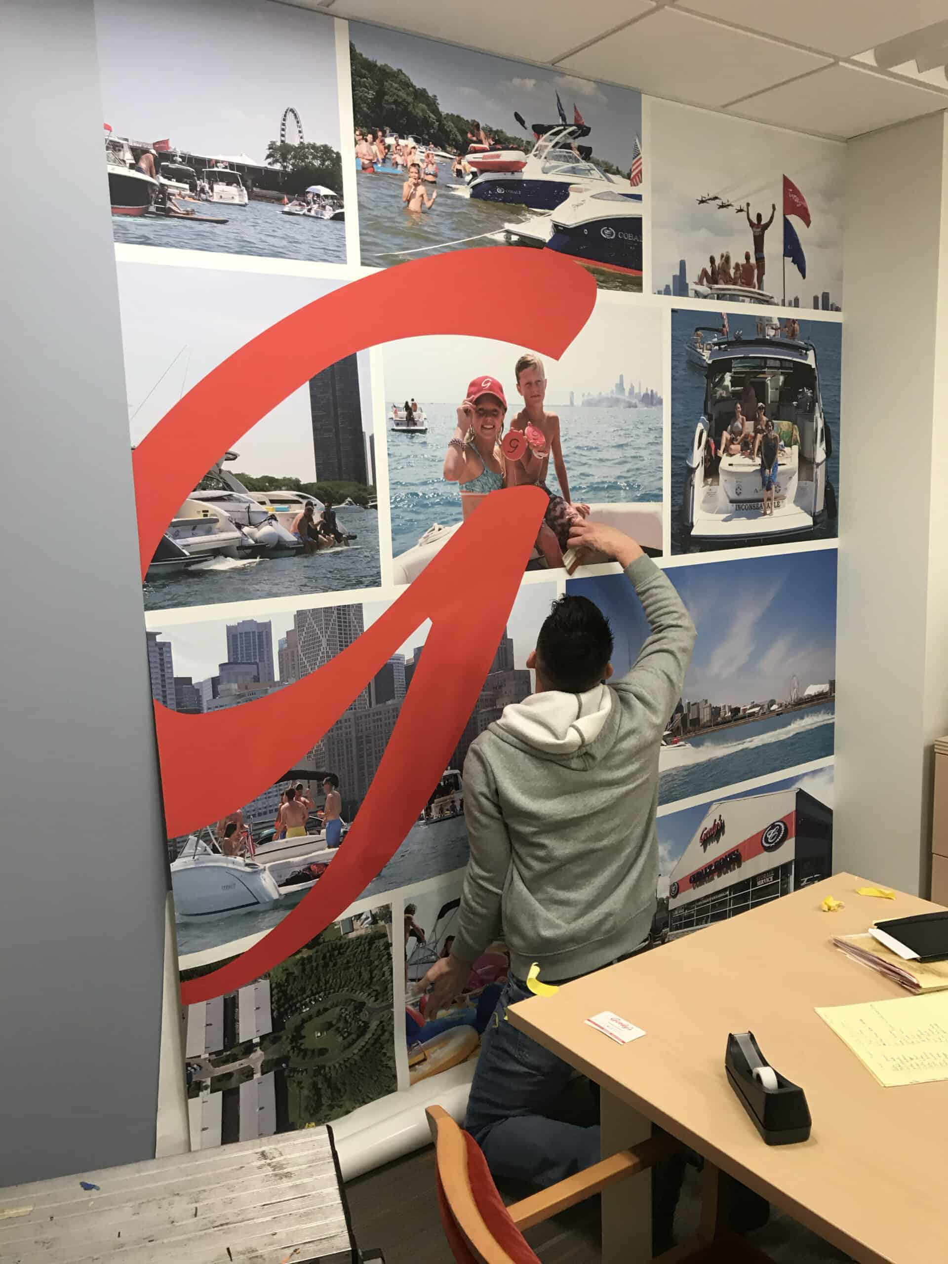 A person in a hoodie installs a wall mural in an office, featuring large boating and water event photos and a bold red letter G across the collage. A desk with office supplies is visible in the foreground.