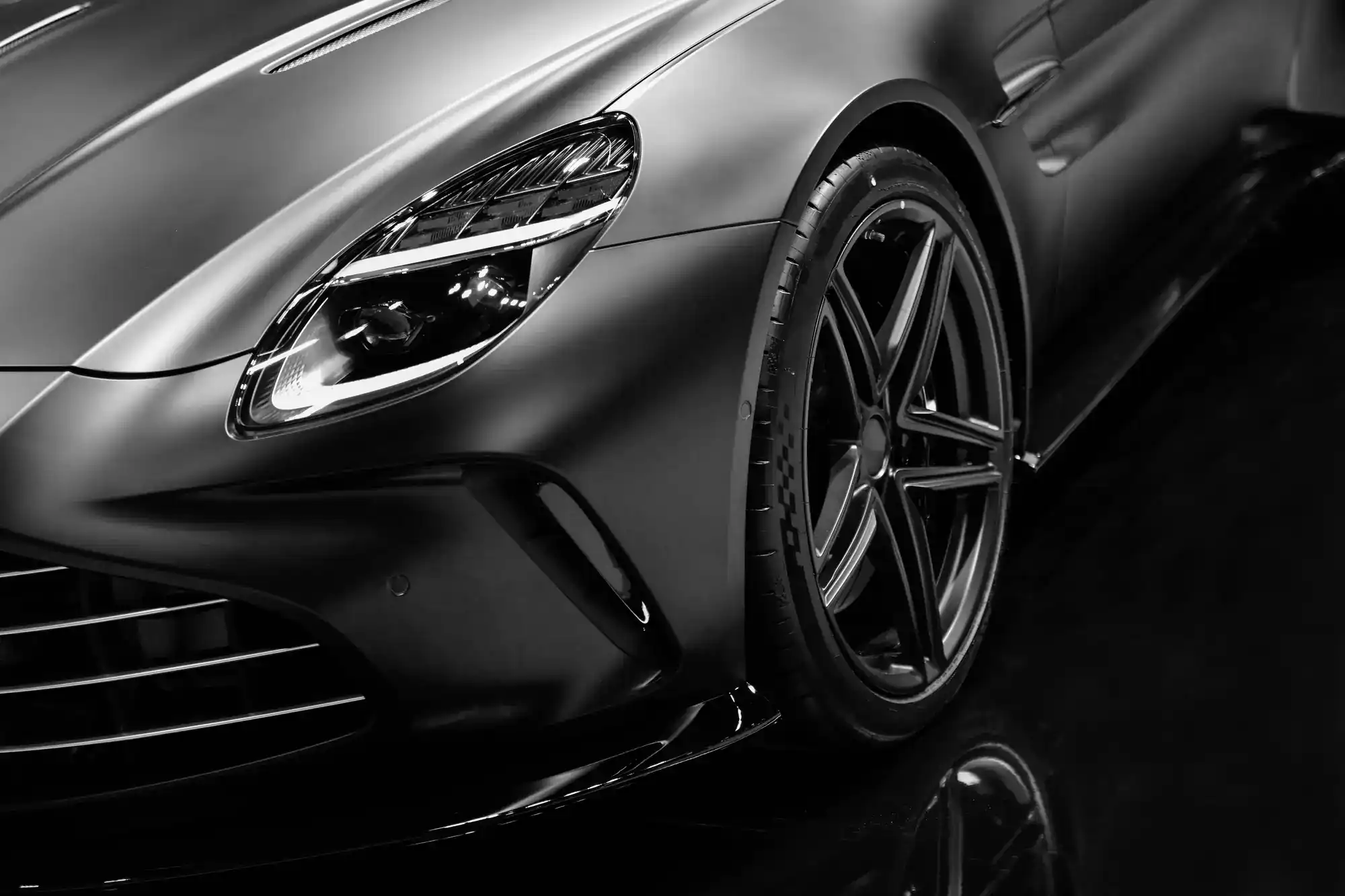Close-up of the front left side of a sleek, matte black sports car, showing the headlight, tire, and aerodynamic body details with a reflective surface underneath.