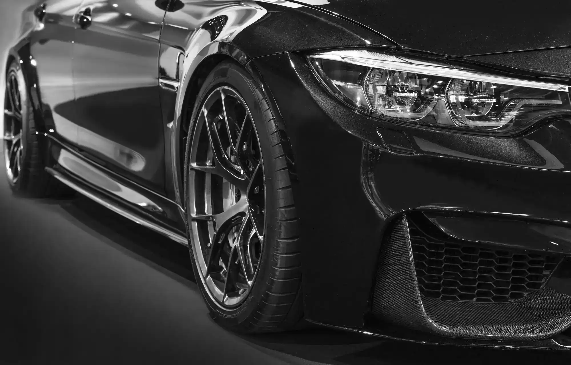 Close-up, black and white photo of the front side of a sleek sports car, highlighting the wheel, tire, headlight, and angled body design. The car has a shiny, reflective surface and modern styling.