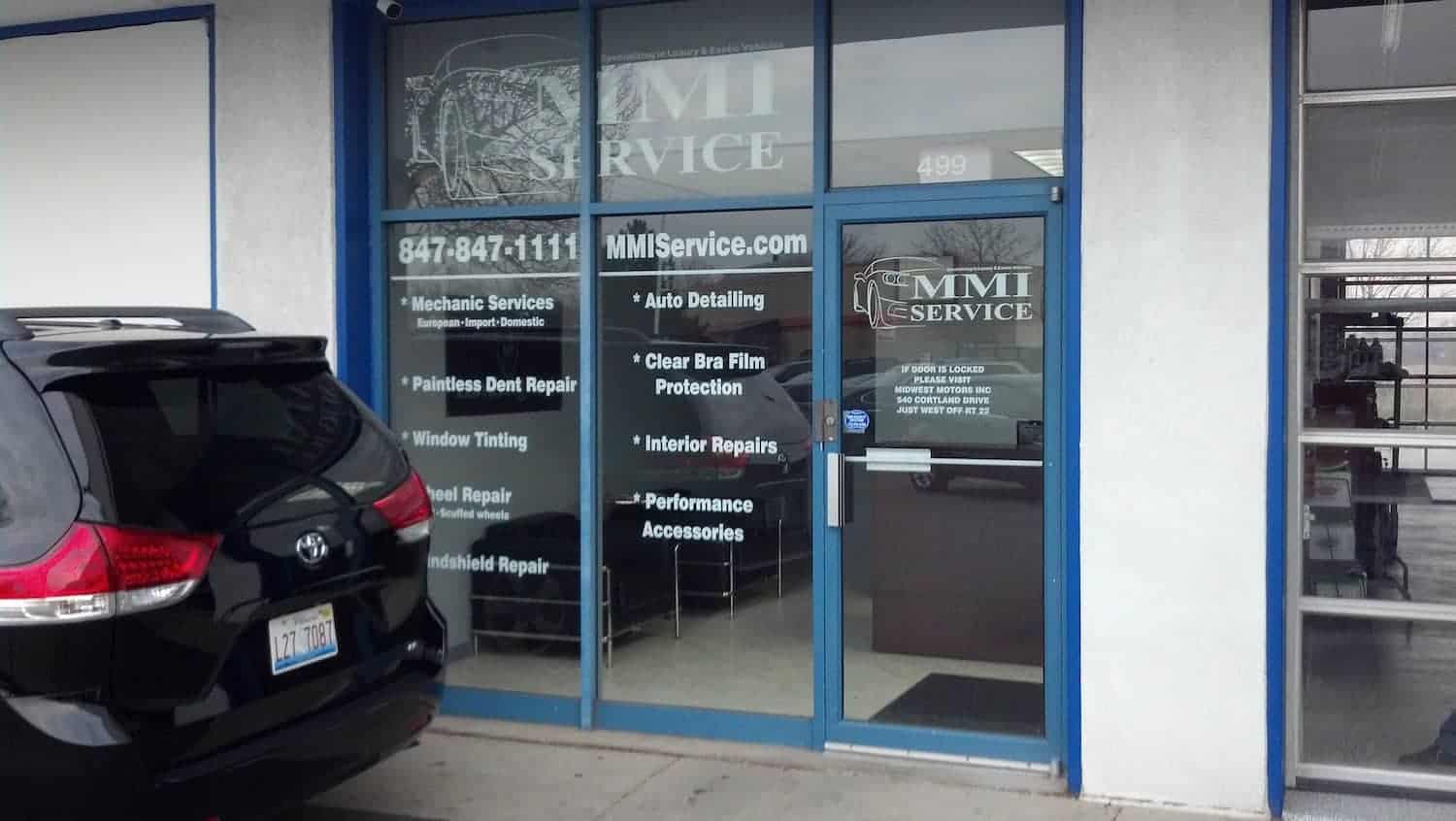 Glass storefront of an auto service shop with "MMI Service" signage and a list of services, including mechanic services, auto detailing, and repairs. A black SUV is parked in front near the entrance.